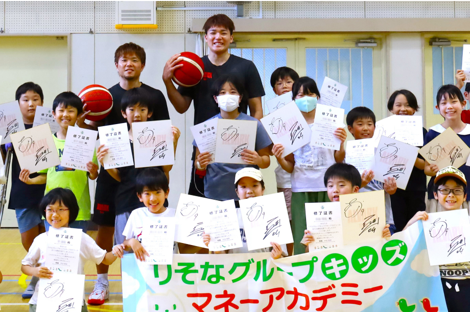 アルバルク東京の岡本選手、安藤選手とキッズマネーアカデミーの修了証書と選手の色紙を持った子供たちの集合写真