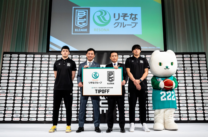 サムネイりそなグループとBリーグのロゴを持った島田チェアマンと南社長を中心に中村選手と三谷選手、りそにゃが並んで記念撮影をしている写真ル